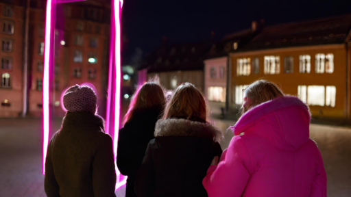 En grupp tjejkompisar står med ryggen mot kameran och tittar på ett neonrosa ljuskonstverk